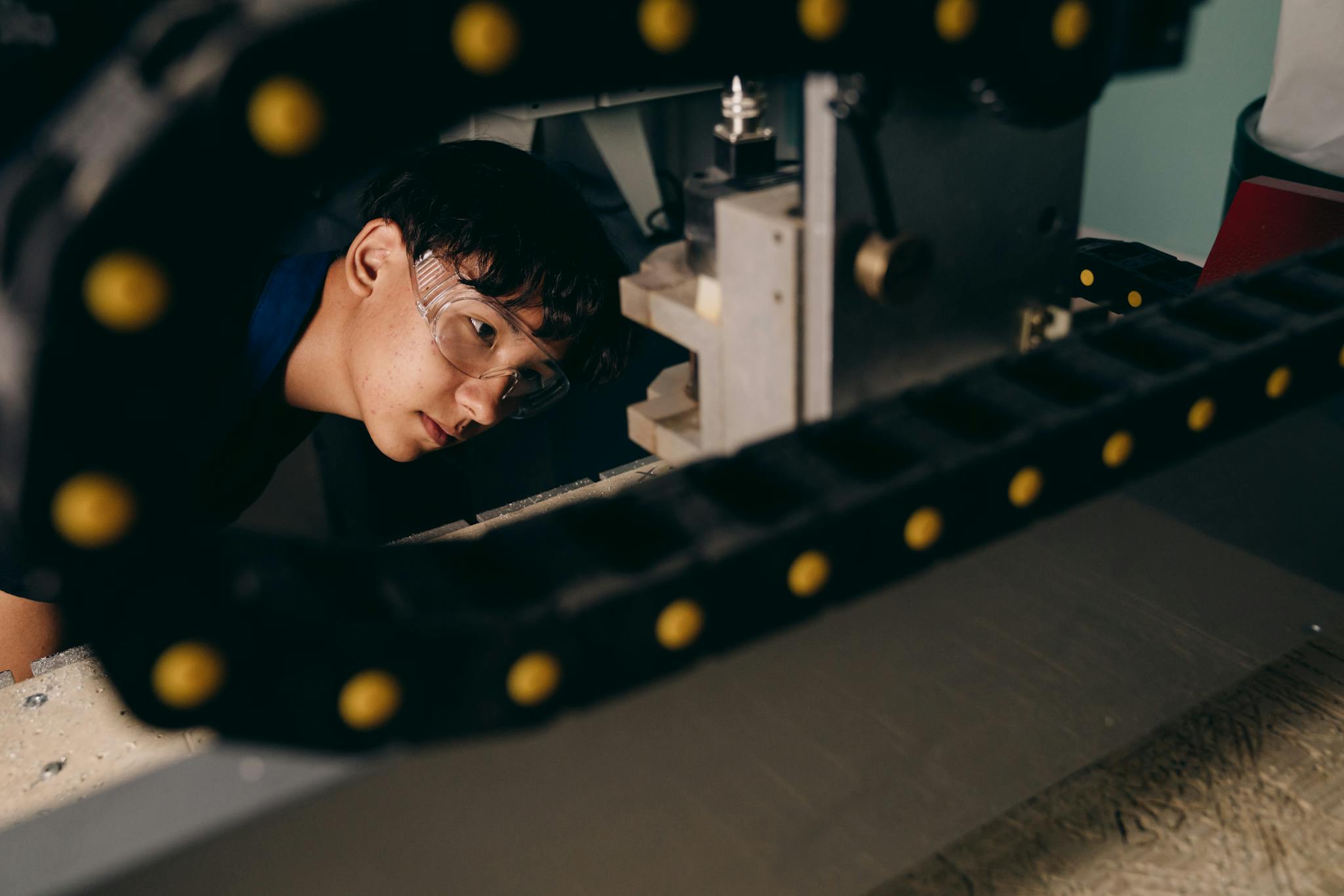 Young Asian engineer wearing goggles, inspecting machinery in a workshop setting.